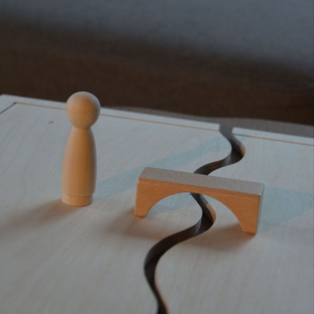 Das Foto zeigt ein Systembrett aus Holz (auch Familienbrett genannt), welches häufig in systemischer Beratung und Therapie verwendet wird. Auf ihm steht eine Holzfigur und ein Baustein in Form einer Brücke.