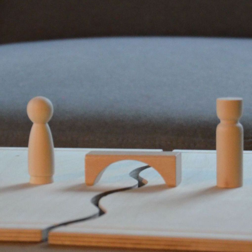 Das Foto zeigt ein Systembrett aus Holz (auch Familienbrett genannt), welches häufig in systemischer Beratung und Therapie verwendet wird. Auf ihm stehen zwei Holzfiguren und zwischen diesen ein Baustein in Form einer Brücke.